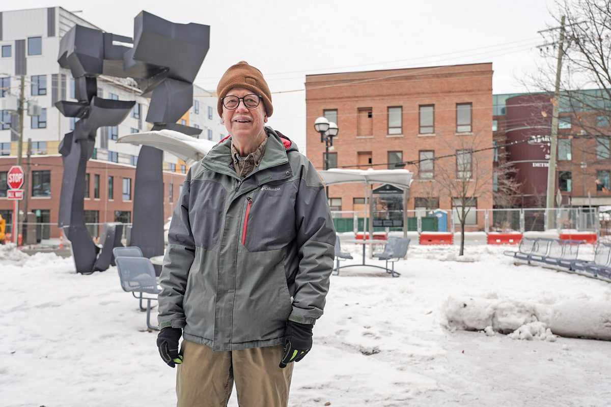 A smiling man in a winter coat, hat, and gloves stands in a wintery city.