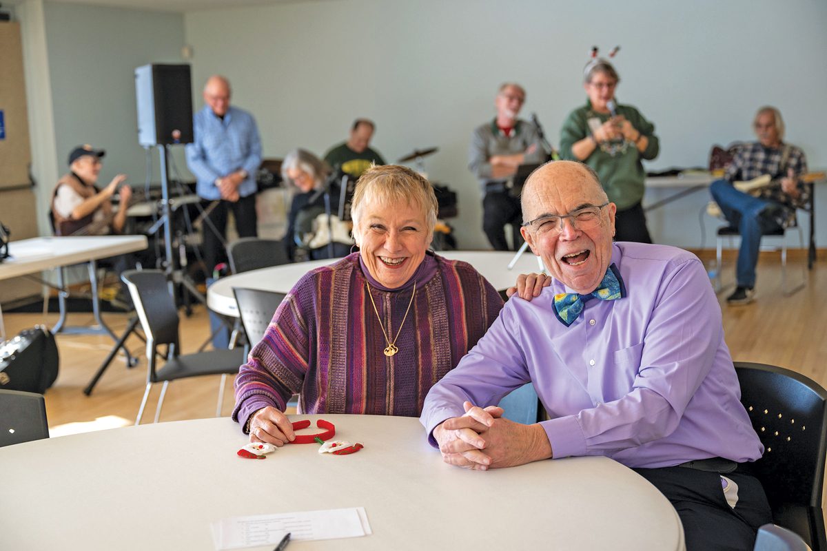 An older woman and an older man sitting in a room and laughing. A band plays behind them. 