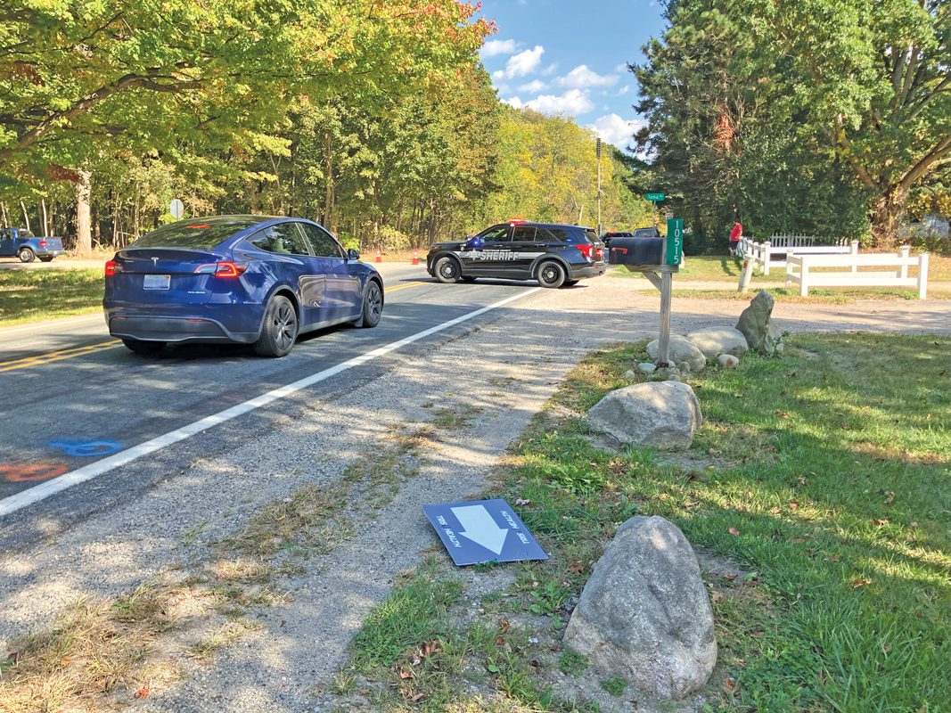 A sheriff's car blocks a road. A sign lays on the ground in the foreground. 