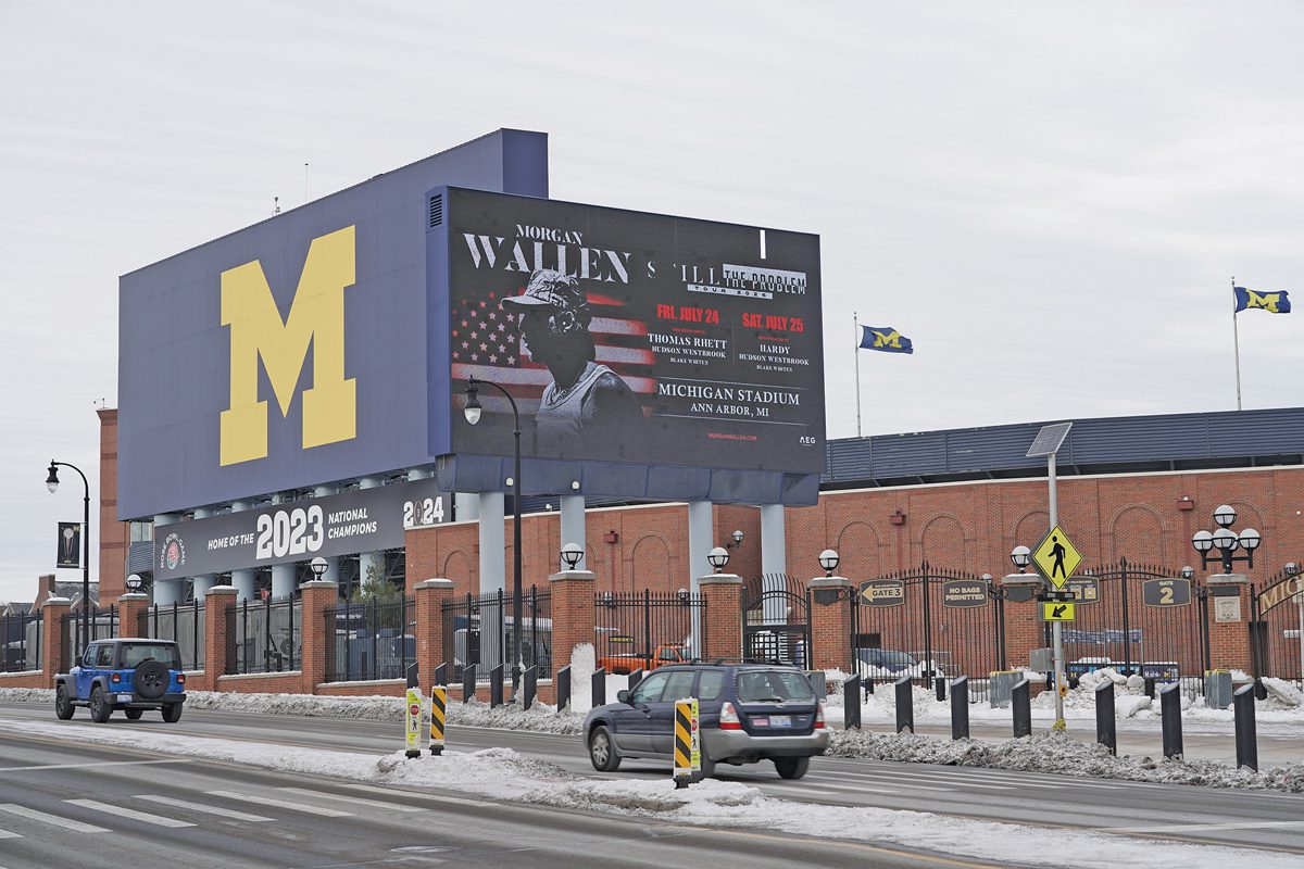 Exterior of Michigan stadium. A large blue sign with a maize-colored block M looms over the stadium. Just in front of it is a large billboard advertising a Morgan Wallen concert.