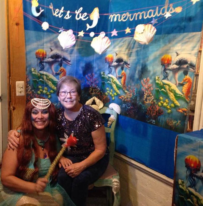 Two women in front of a banner with an undersea theme and the words "let's be mermaids." One of the women is dressed as a mermaid.