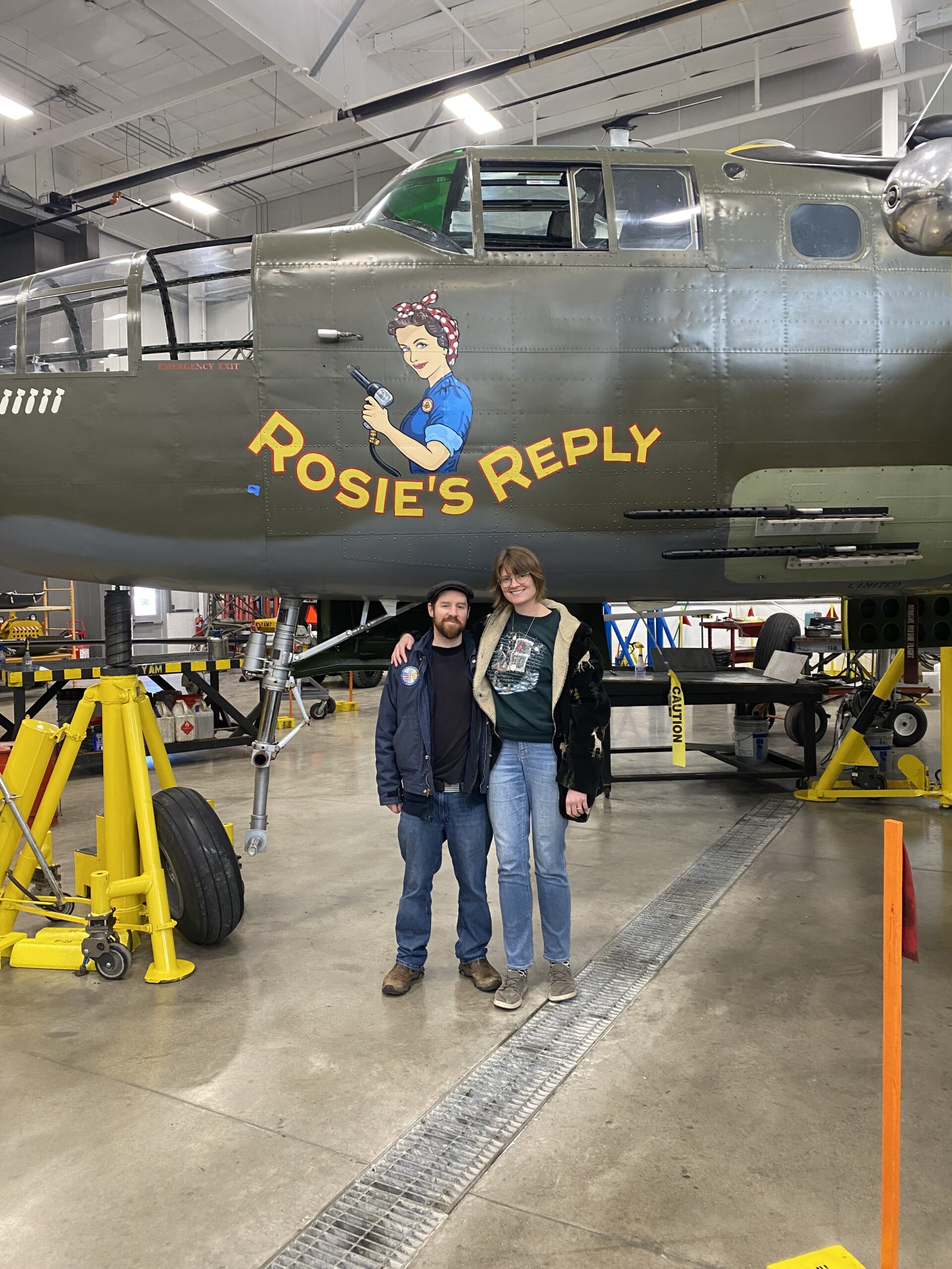 Doug and Brooke standing in an airplane hanger in front of a B-24 plane called Rosie's Reply.