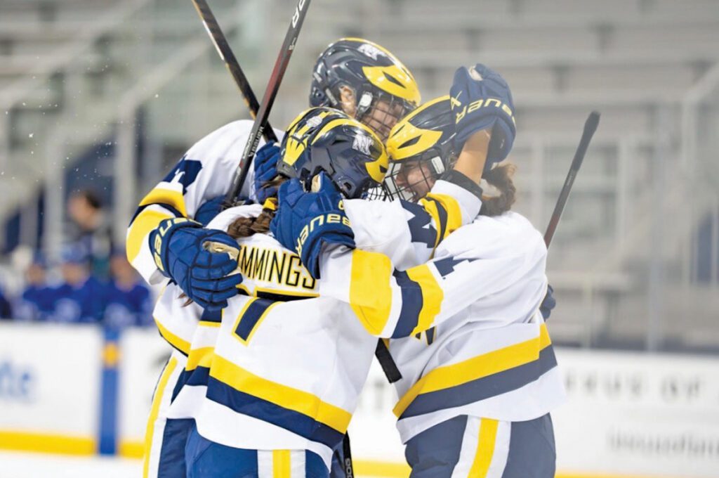 Three women's hockey players in hockey gear hug on the ice.