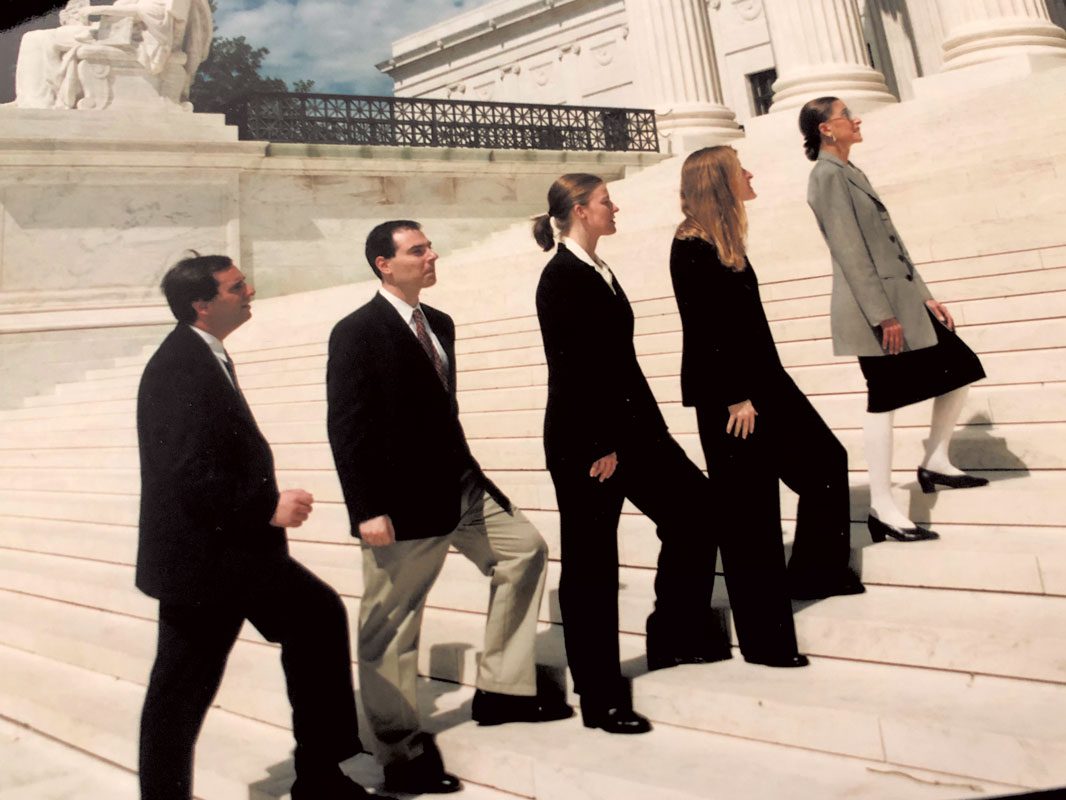 Four clerks in suits stand behind Ruth Bader Ginsburg on the Supreme Court steps. 