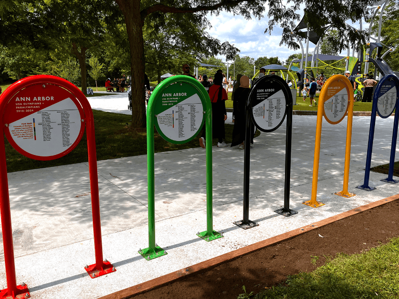 A row of colorful circular signs recognizing Ann Arbor Olympians and Paralympians lines a paved pathway in a public park. An accessible playground, mature trees and visitors enjoying the park are visible in the background.