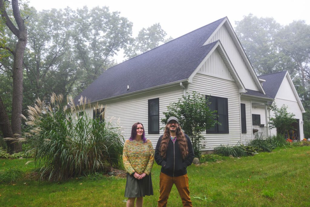 Michelle and Mike Balhorn stand in front of their white house in the woods on a cloudy day.