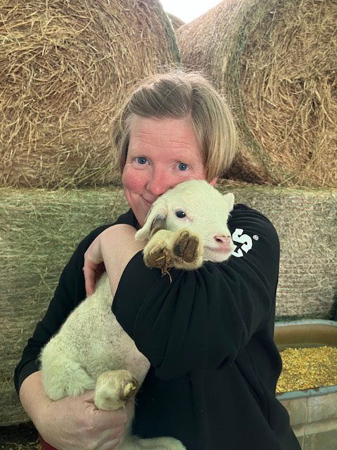 Sarah Schloss holds a lamb while standing in a field.
